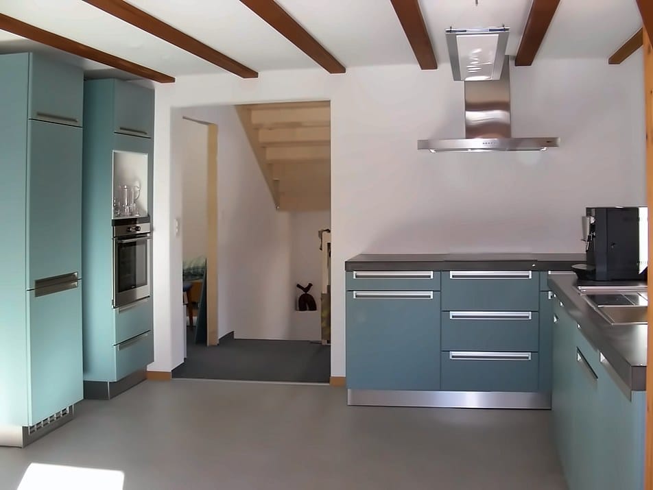 Offene Küche mit petrolblauen Fronten, Dunstabzug und Holzbalkendecke, links die Treppe