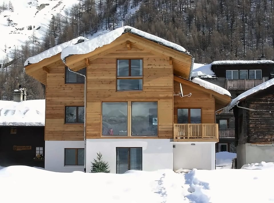 Sonnenbeschienene Holzfassade des Chalets mit grossen Fenstern und Balkon, Dach und Boden tief verschneit